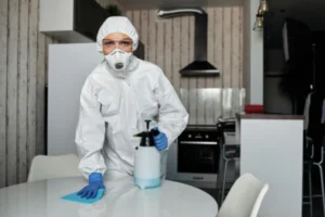 A professional cleaner in a white hazmat suit, mask, and blue gloves wiping down a kitchen table with a spray bottle.